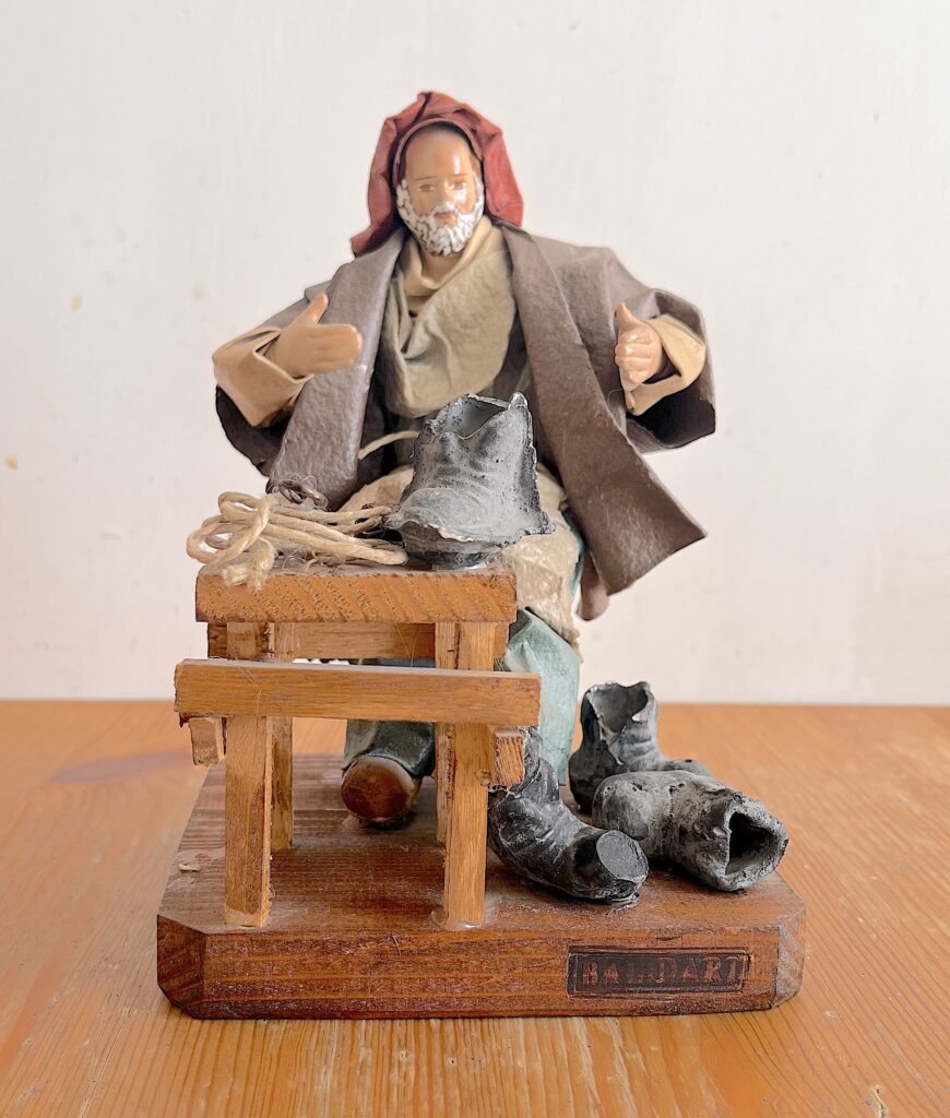 Figura artigianale vintage in cartapesta leccese raffigurante un uomo barbuto seduto davanti a un banco da lavoro in legno, con abiti in tessuto.