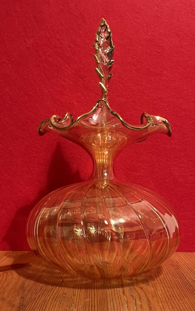 Bottiglia decorativa in vetro soffiato color ambra, con corpo arrotondato a coste, bordo ondulato dorato e decorazione superiore fiammeggiante.