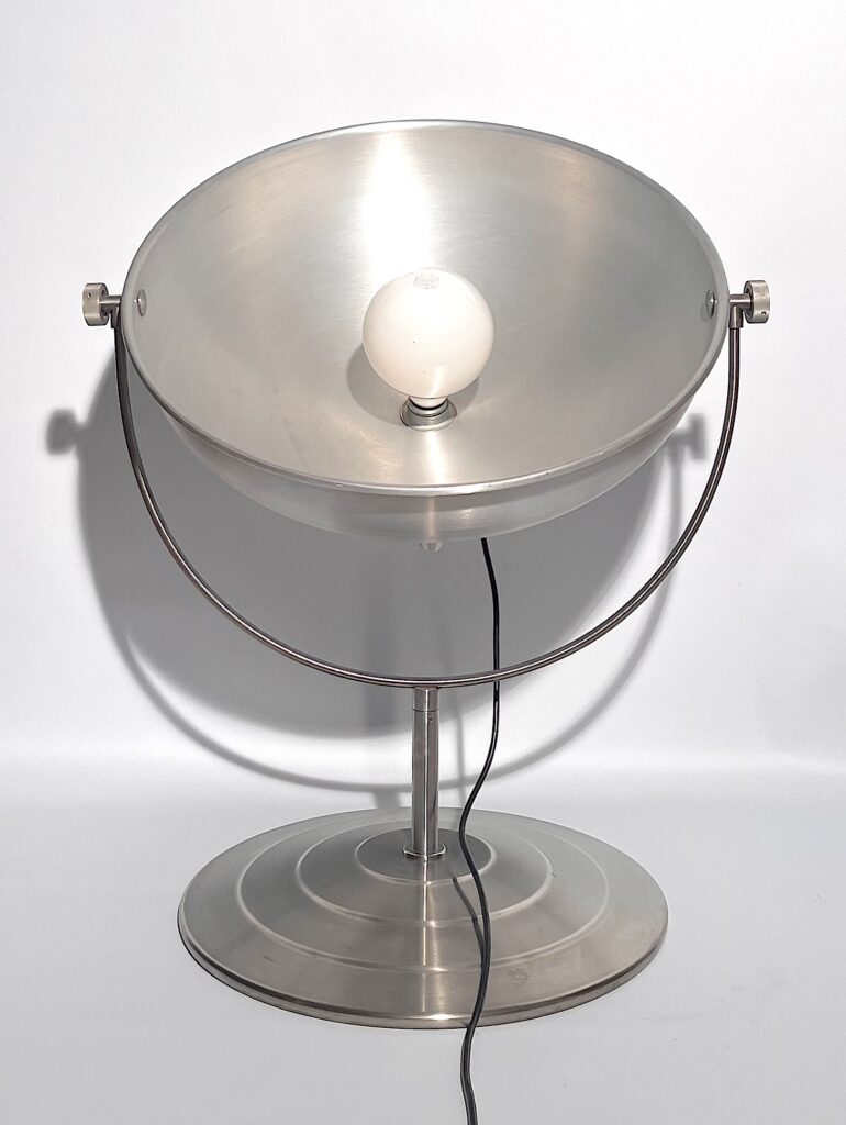 Lampada da terra in metallo argentato con grande riflettore circolare orientabile, sostegno ad arco e base rotonda con cerchi concentrici.